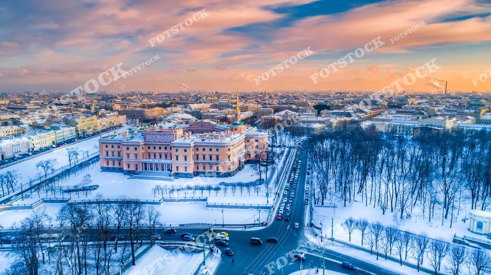 Фотография зимнего Санкт-Петербург, Россия, сделанная на закате солнца. На изображении показана панорама города с высоты Михайловский замок (Инженерного замка). Фотоснимок передаёт атмосферу зимнего Петербурга: тёплые закатные оттенки, архитектурный силуэт замка и улицы исторического центра города. Фото подчёркивает особенности петербургской архитектуры и выразительные городские виды северной столицы. Иллюстрация может использоваться как изображение, картинка или слайд, посвящённые архитектуре и панорамам российских городов.